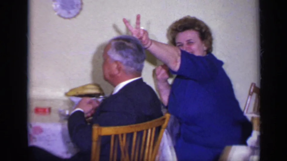 A woman in blue leans forward laughing with two fingers raised behind the man at a private California dining table.