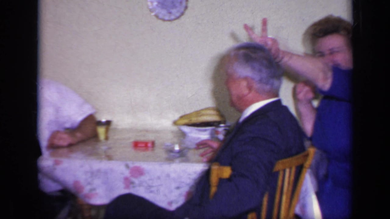 1969 California home-movie frame showing adults at a covered table, with a woman in blue making a peace sign behind an older man.