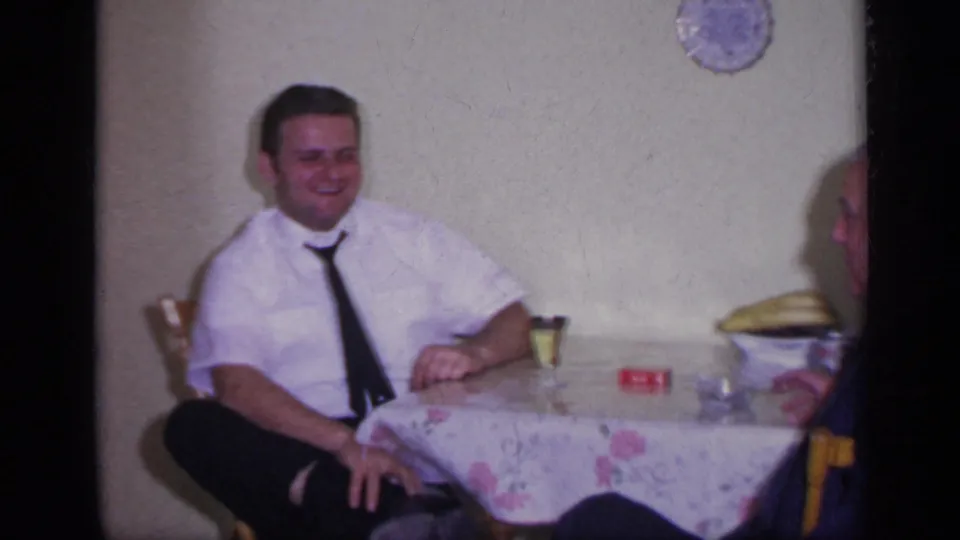 1969 California home-movie frame of a smiling man in a white shirt and dark tie seated beside a floral tablecloth.