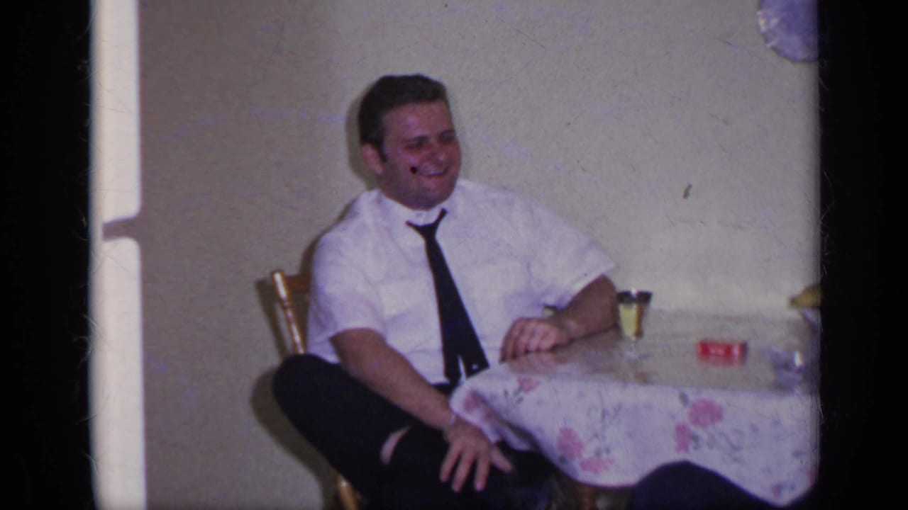 1969 California home-movie frame showing a seated man smiling beside a table with a floral cloth and small glass.