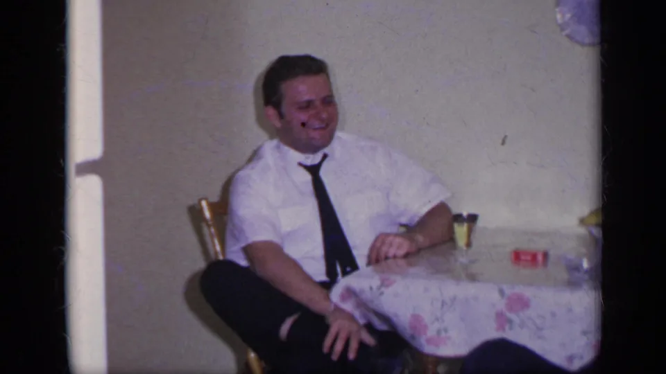 1969 California home-movie frame showing a seated man smiling beside a table with a floral cloth and small glass.