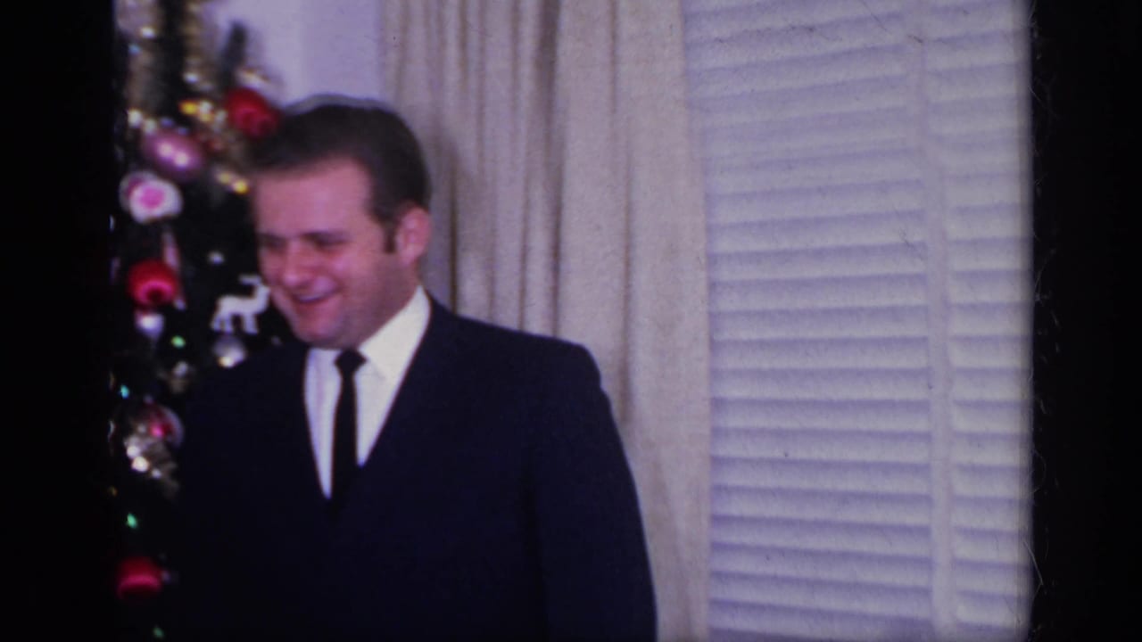 1969 California home-movie frame of a man in a dark suit standing near a decorated Christmas tree inside a home.