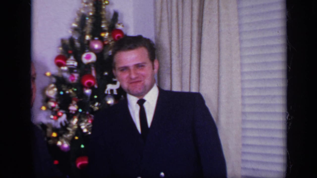 Younger man in a dark suit standing beside a decorated Christmas tree indoors.