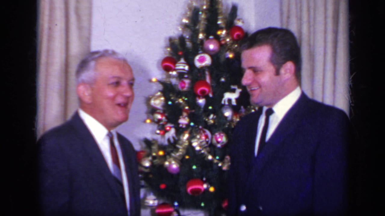 Older and younger men in suits smiling near a decorated Christmas tree.