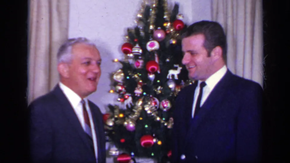Two adult men in suits stand in a private California interior beside a decorated Christmas tree in this 1969 home-movie frame.