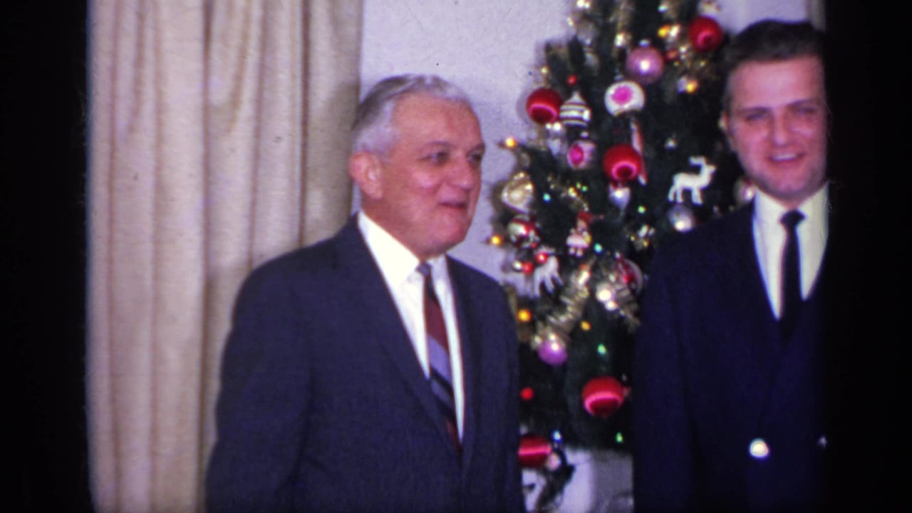 A 1969 California home-movie frame shows two suited adult men near a Christmas tree, with curtains and holiday decorations visible.