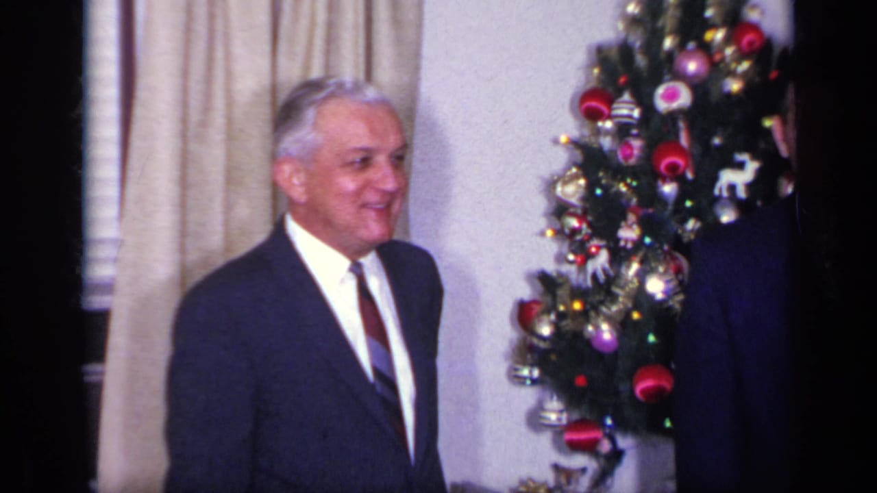 White-haired man in a suit smiles beside a decorated Christmas tree indoors.