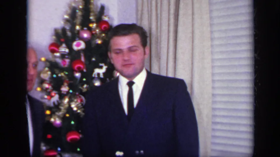 A younger man in a dark suit and black tie stands near a brightly decorated Christmas tree in the 1969 California home movie.