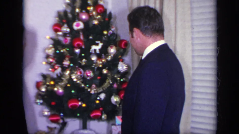 The younger suited man turns toward the Christmas tree and window blinds inside a private 1969 California holiday setting.