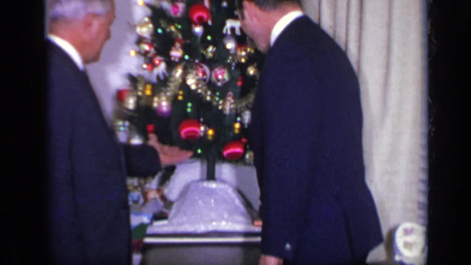 In a 1969 California home interior, two men in dark suits stand beside a decorated Christmas tree with red ornaments and curtains behind them.