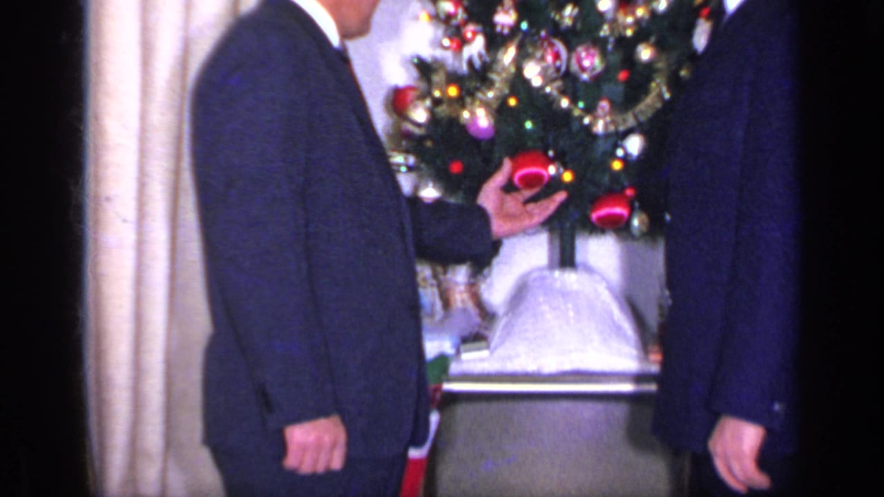 A suited man gestures near a decorated Christmas tree as another man stands at the edge of frame.