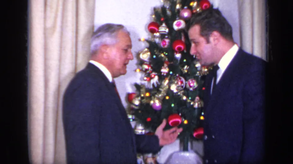 Two men in dark suits converse beside a lit Christmas tree with red and metallic ornaments in a 1969 California home interior.