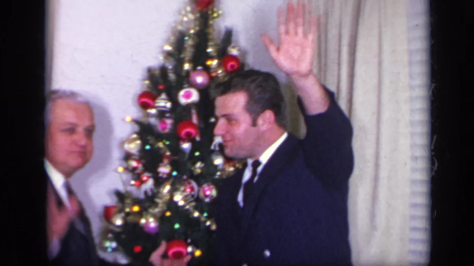 A man in a dark suit raises his hand toward the camera beside the Christmas tree, with another suited man partly visible in the 1969 California interior.