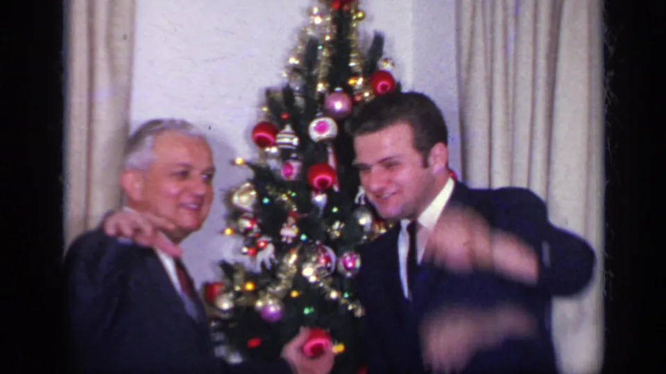In a 1969 California home movie frame, two men in dark suits pose by a decorated Christmas tree as one waves toward the camera.