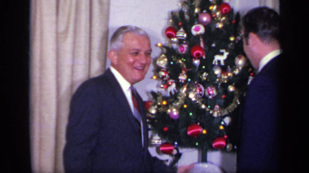 An older man smiles near a decorated Christmas tree while another man is seen from behind.