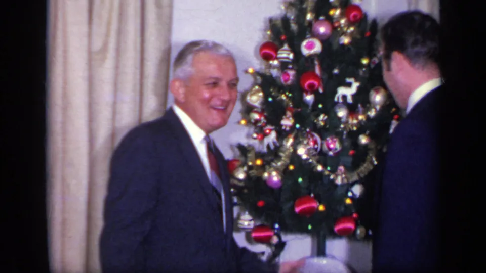An older suited man smiles near the Christmas tree in a California home interior, with a younger man turned away in the foreground.