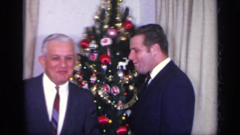Two men in suits stand near the lit Christmas tree, with the older man facing the camera and the younger man turned in profile.