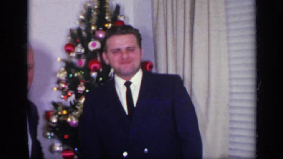 A younger man in a dark double-breasted suit stands near the Christmas tree, with curtains and a window blind behind him.
