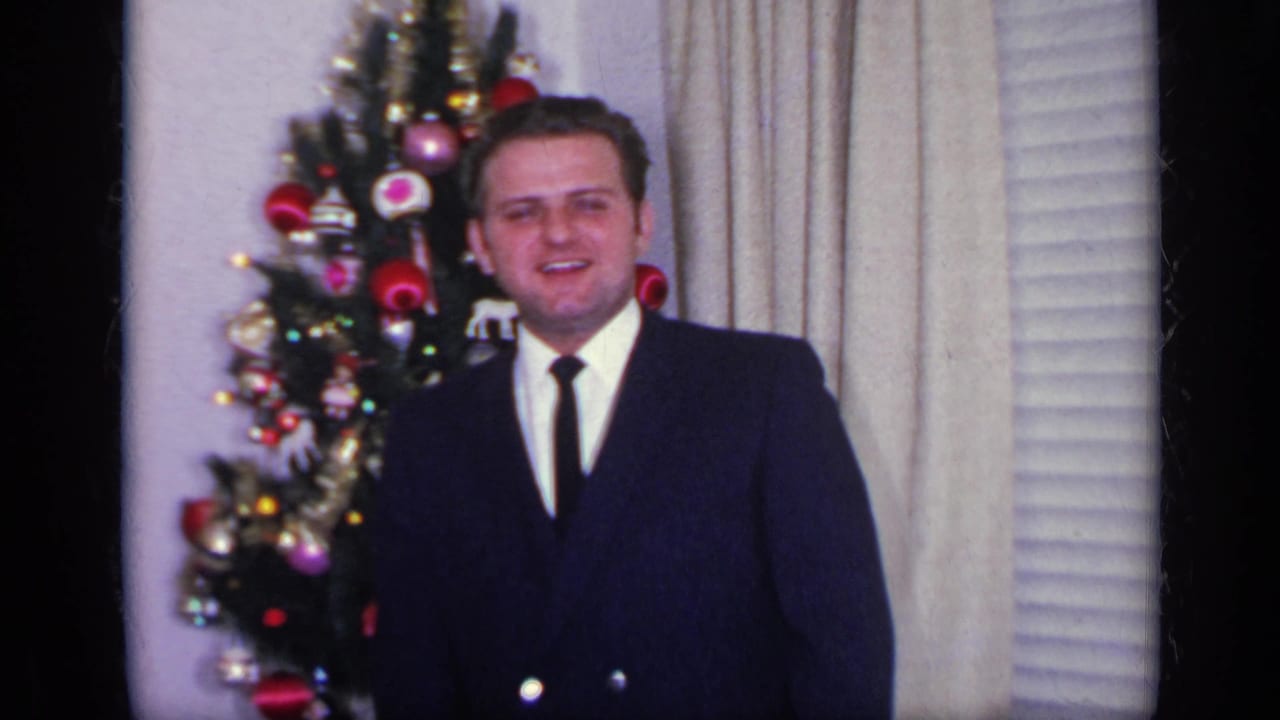 Man in a dark suit standing beside a small decorated Christmas tree indoors.