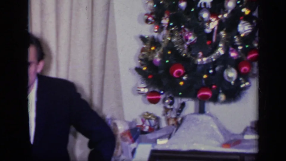 A suited man is partly cut off at the edge while the home movie frames a decorated Christmas tree in the living room.