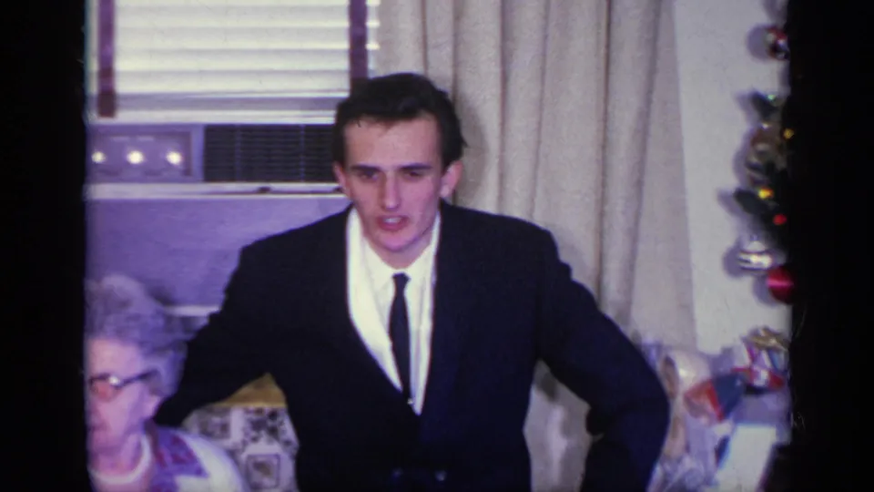 A suited man leans forward in a 1969 California living room as a seated woman and decorated Christmas tree appear nearby.