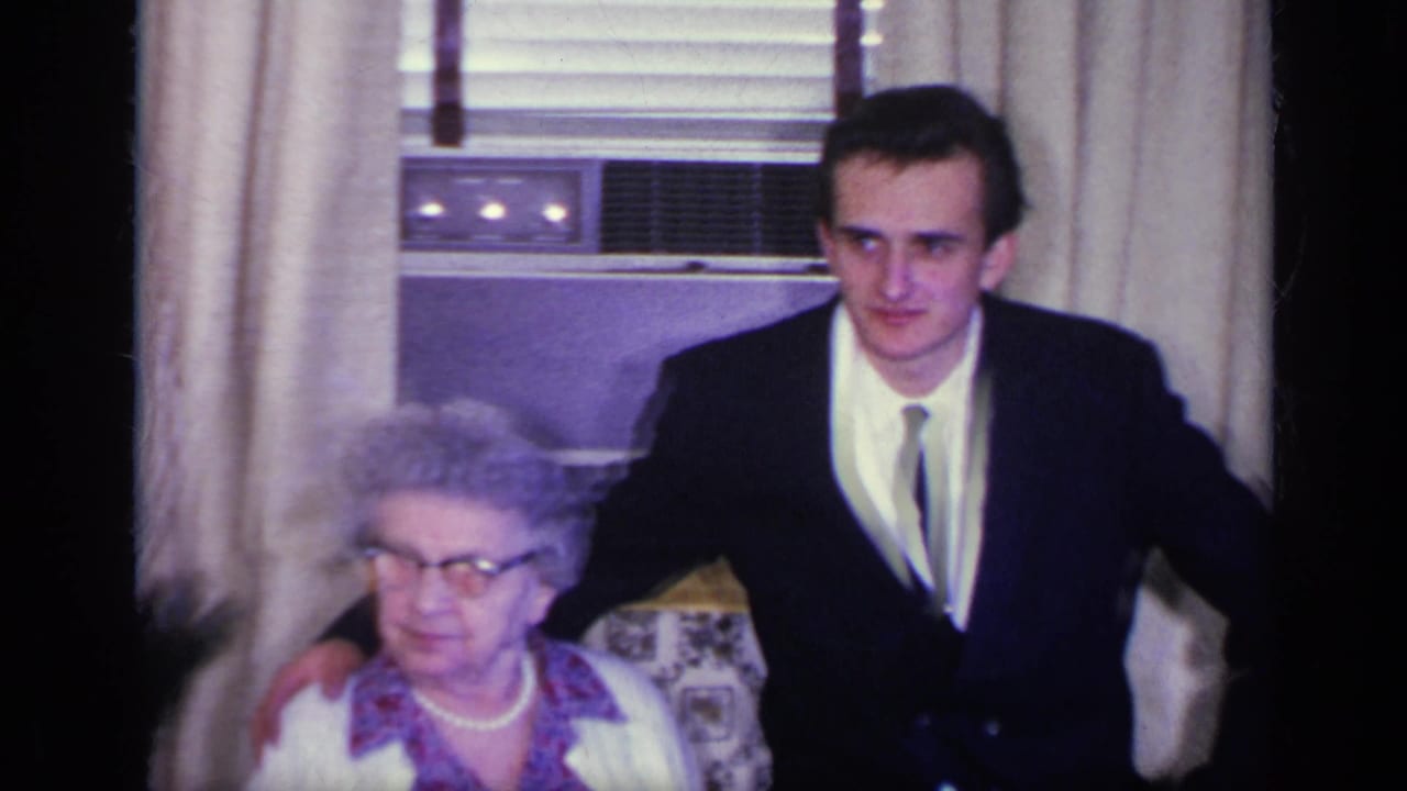 Young man in a dark suit stands beside an elderly woman seated in a patterned armchair.