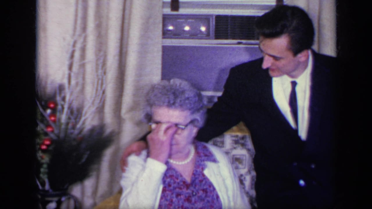 Elderly woman touches her glasses while a suited young man stands beside her chair.