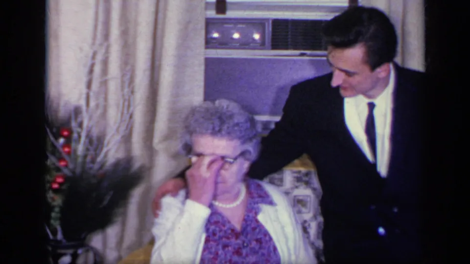The elderly woman adjusts her glasses as the young man in a dark suit leans beside her in the living room.