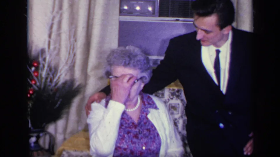 A suited young man stands beside the seated woman, who covers part of her face in an emotional-looking moment.