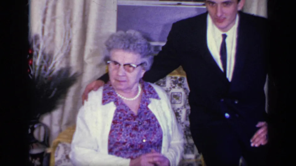 The woman sits with hands lowered as the suited young man stands close beside her in the 1969 California living room.