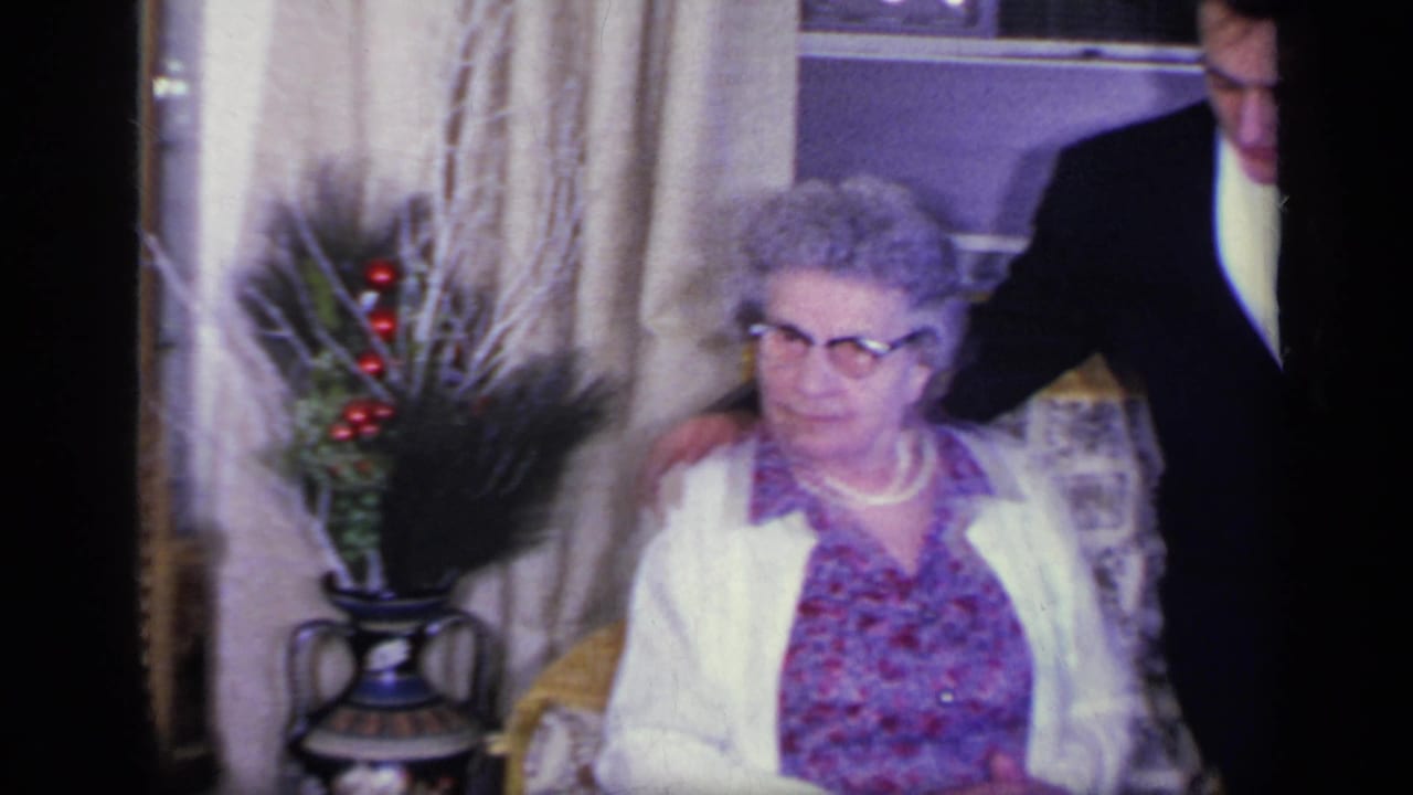 Elderly woman in a floral dress and white cardigan seated beside formally dressed guests in a living room.