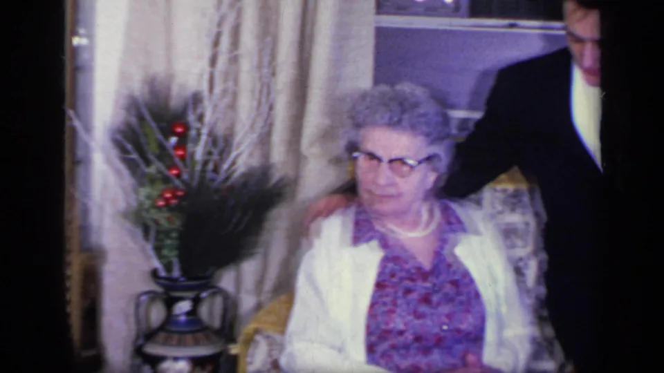 1969 California home-movie frame of an elderly woman seated near suited guests, with curtains, a gold-framed mirror, and a decorative vase behind her.