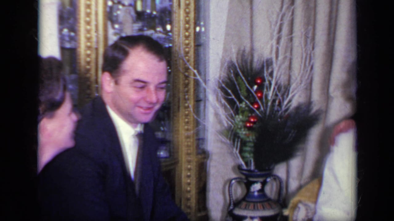 Man in a dark suit smiles while seated beside another guest in a decorated living room.