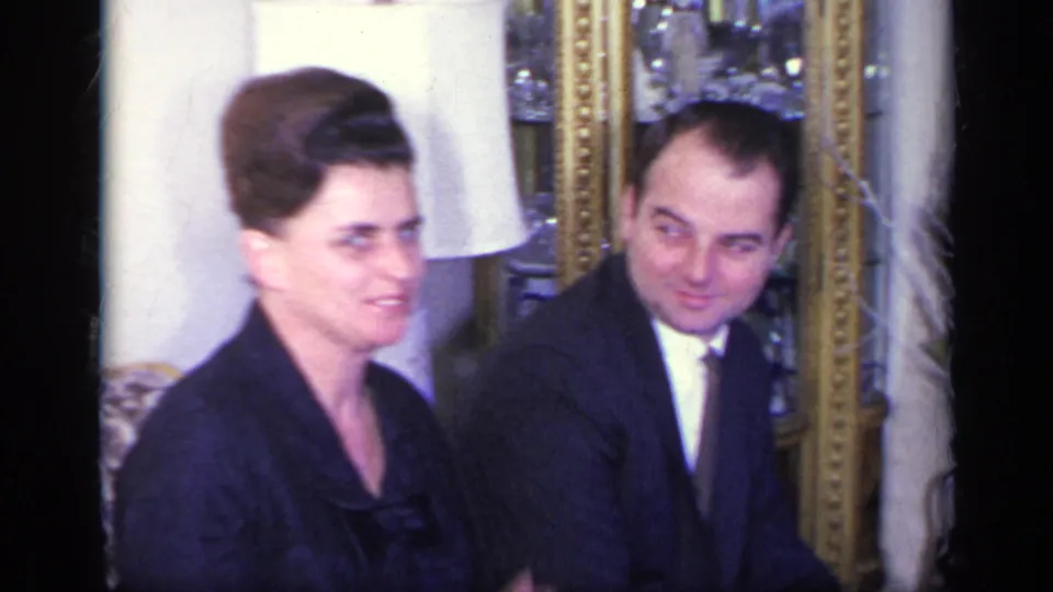 1969 California home-movie frame of a formally dressed woman and suited man seated together in a living room with a lamp and framed mirror.