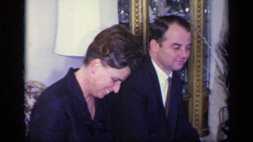 1969 California home-movie frame of a woman smiling downward next to a suited man, with formal living-room decor visible behind them.