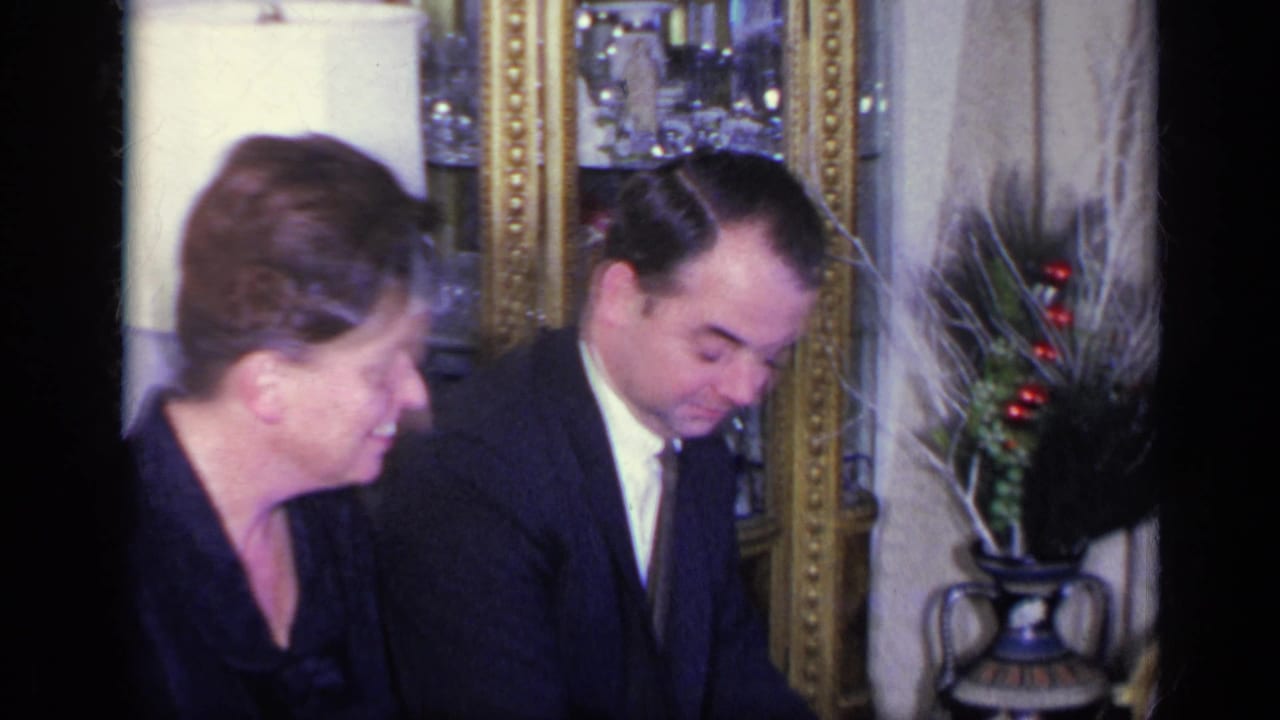 Woman in black dress sits beside a man in a dark suit near a gold-framed mirror.