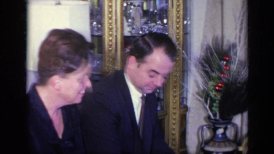 1969 California 8mm frame showing a woman in a black dress and a suited man seated in an ornate private interior.