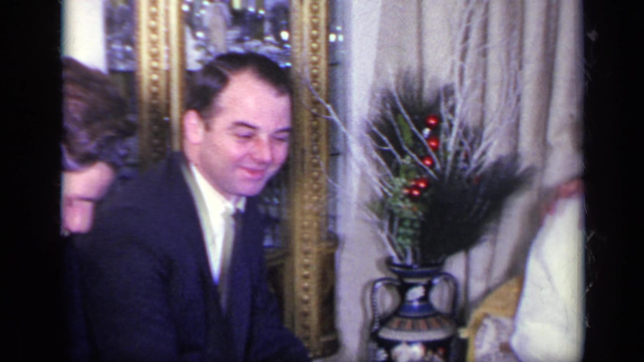 Man in dark suit smiles near a mirror and decorative vase with red berries.