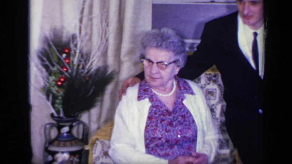 1969 California frame showing an elderly woman in glasses and white cardigan seated while a suited man stands at her side.