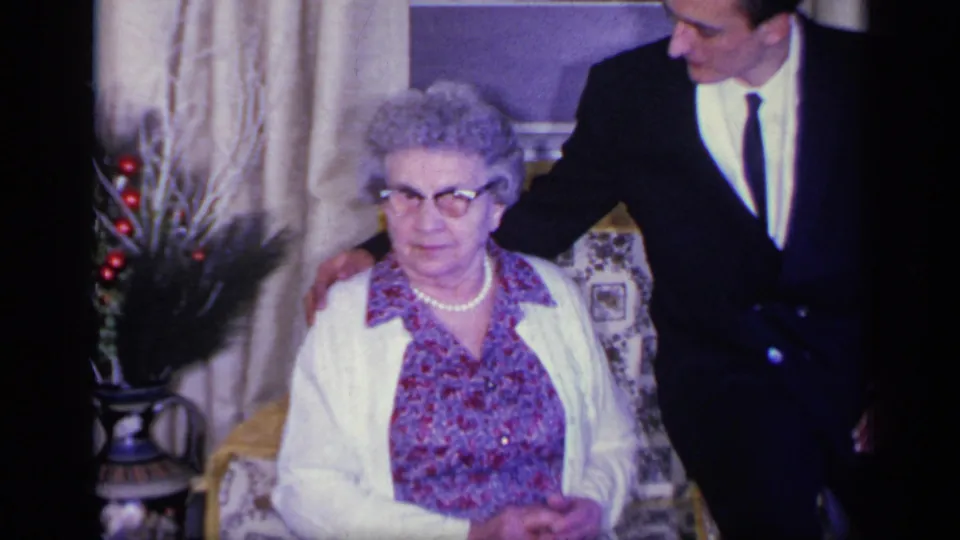 1969 California home-movie frame of an elderly woman seated in a private interior with a suited man standing beside her.