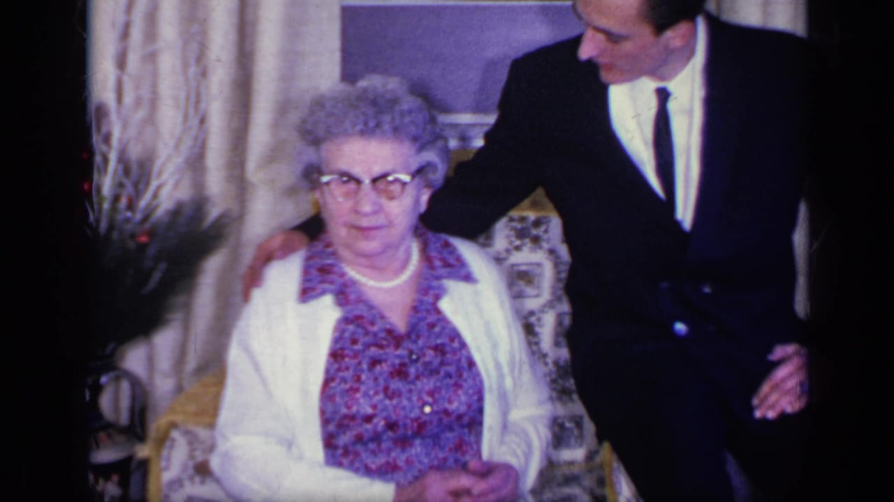 Elderly woman seated in a floral armchair as a young man in a dark suit stands beside her.