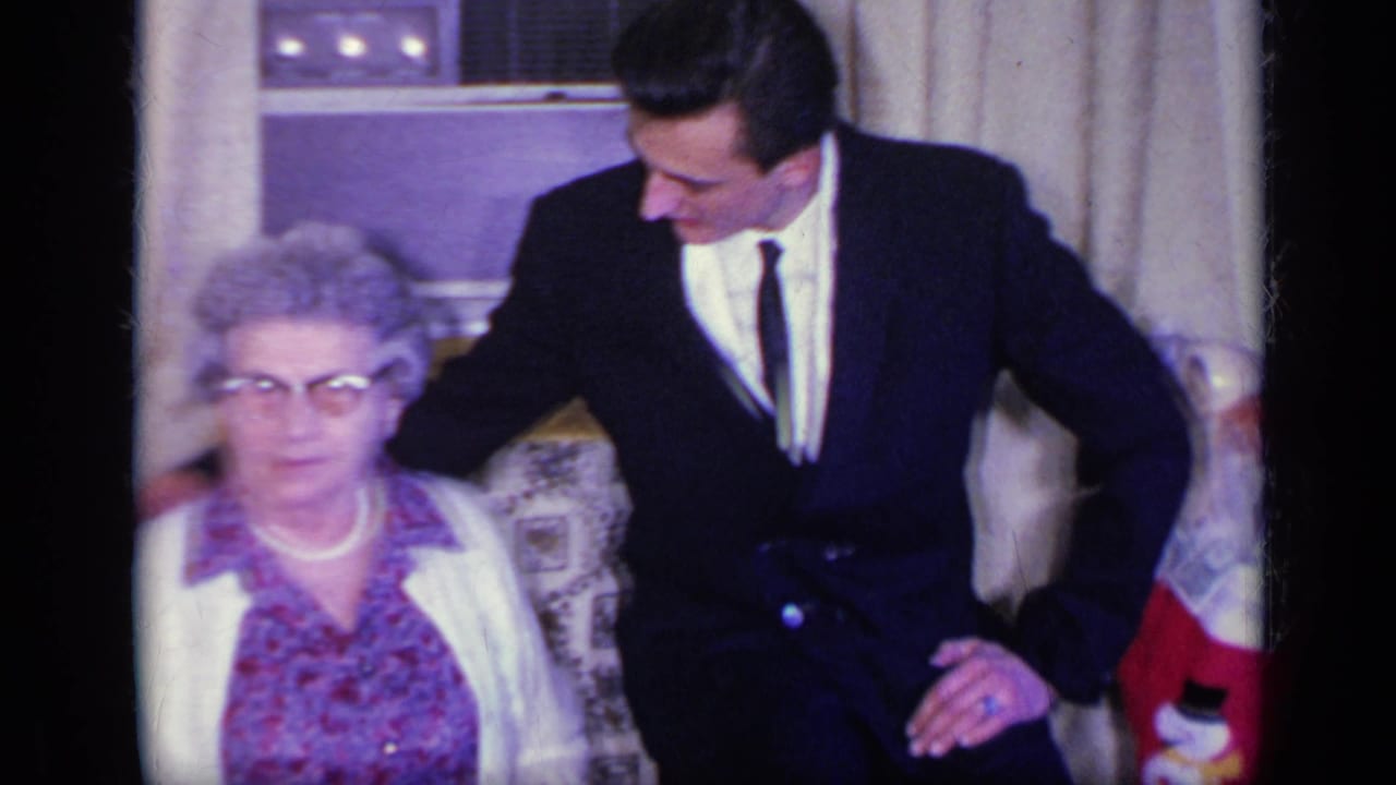 Young man in a dark suit leans beside an elderly woman seated near curtains and a window unit.