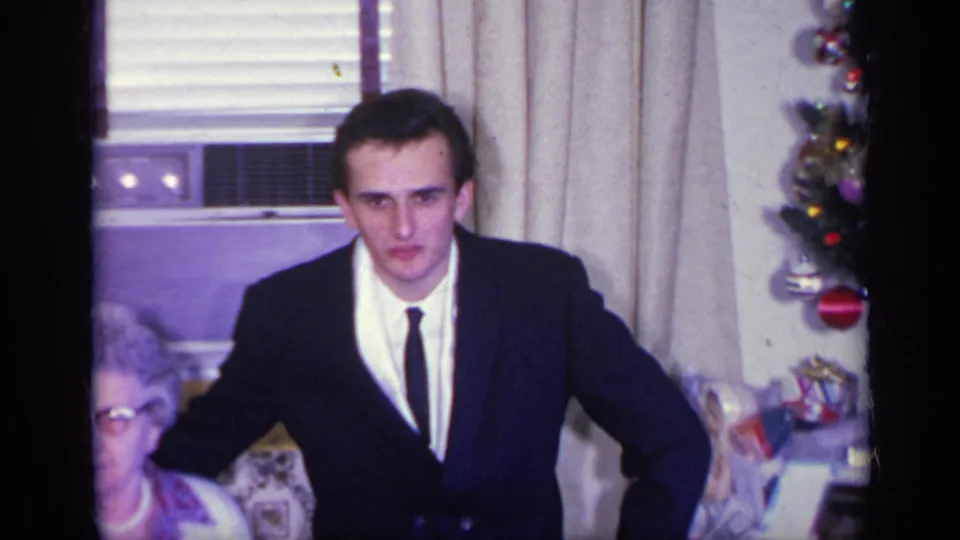 1969 California home interior with a young man in a suit facing the camera, the elderly woman partly cropped, and holiday decor nearby.