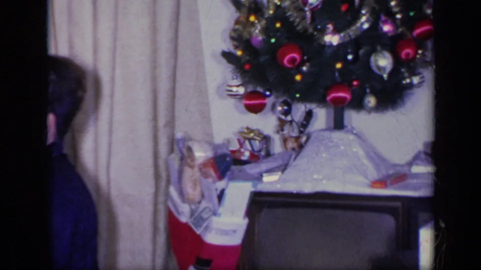 1969 California home-movie frame showing a tabletop Christmas tree with ornaments, tinsel, stockings, and small gifts inside a living room.