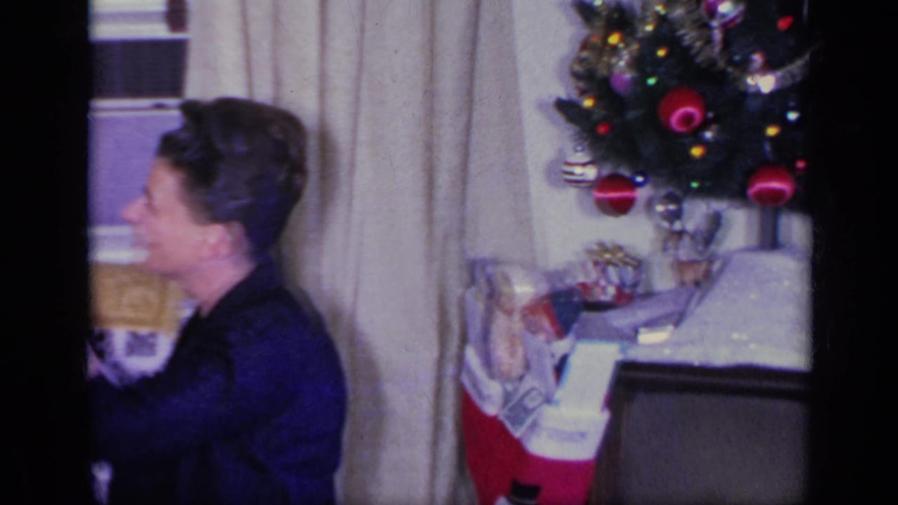 Woman in dark clothing beside a Christmas tree and stocking in a curtained living room.