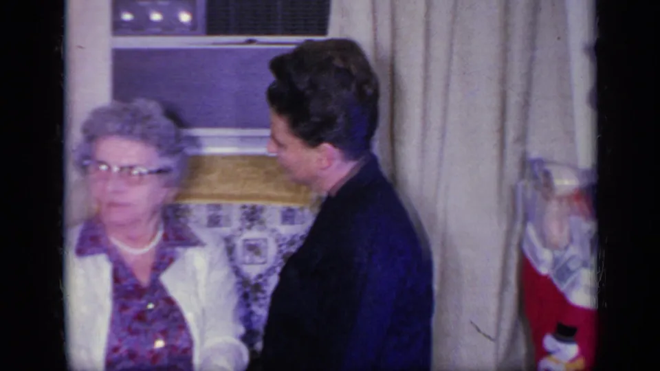 An elderly woman with glasses sits in a living room while a younger woman stands close beside her during a 1969 California Christmas gathering.