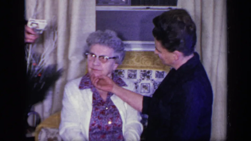 A younger woman reaches toward an elderly woman's collar as they sit and stand near holiday decorations in a 1969 California home interior.