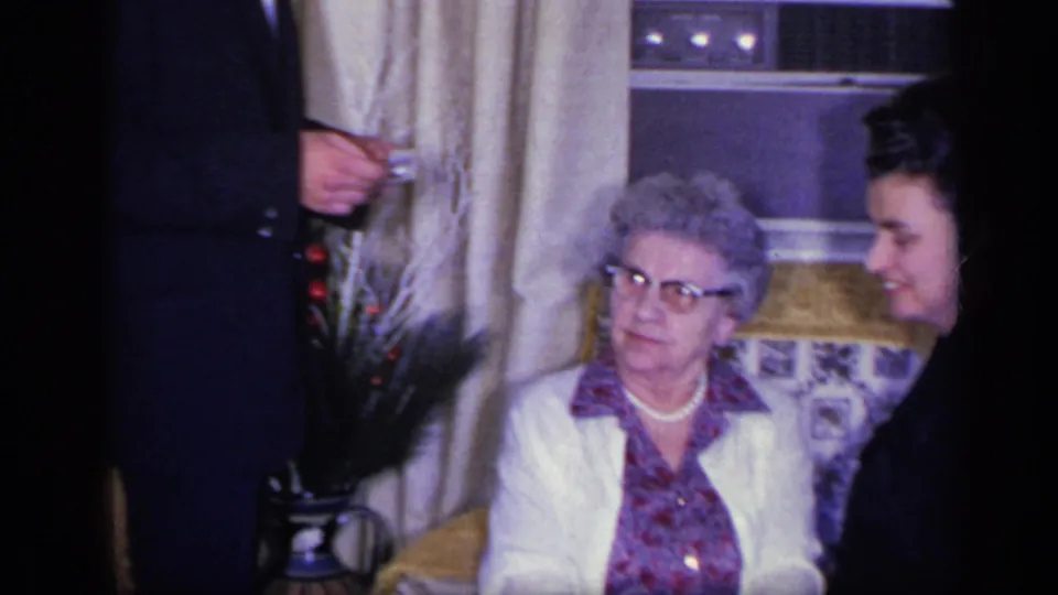 The elderly woman turns toward a smiling younger woman in a private California living room with Christmas decor, curtains, and upholstered seating.