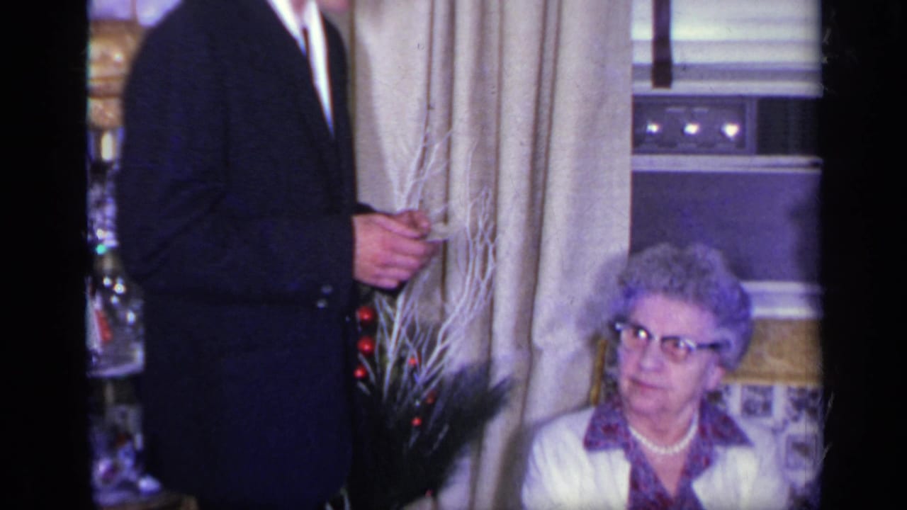 Young man in a dark suit stands beside an older seated woman in a living room.