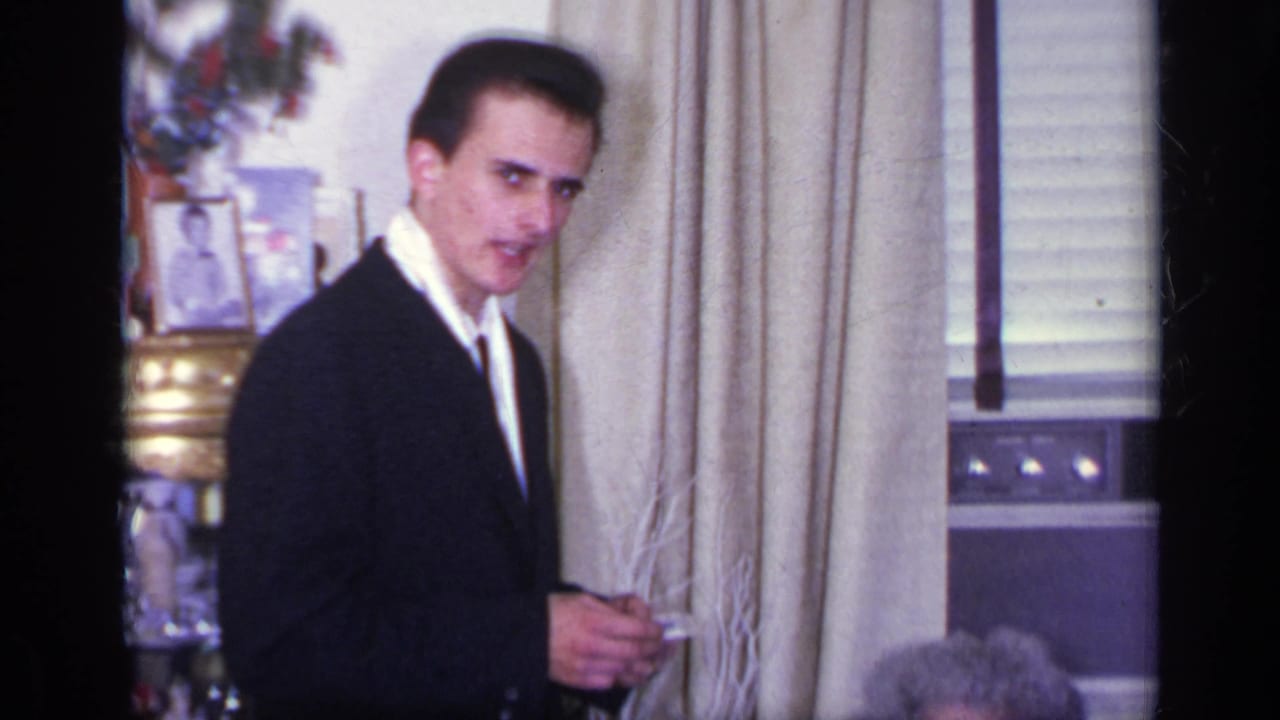 Suited young man looks toward the camera near curtains and a window.